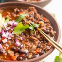 Two bowls of beef chili with a gold spoon, sprinkled with red onions and cilantro.