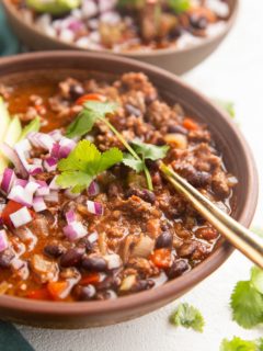 Two bowls of beef chili with a gold spoon, sprinkled with red onions and cilantro.