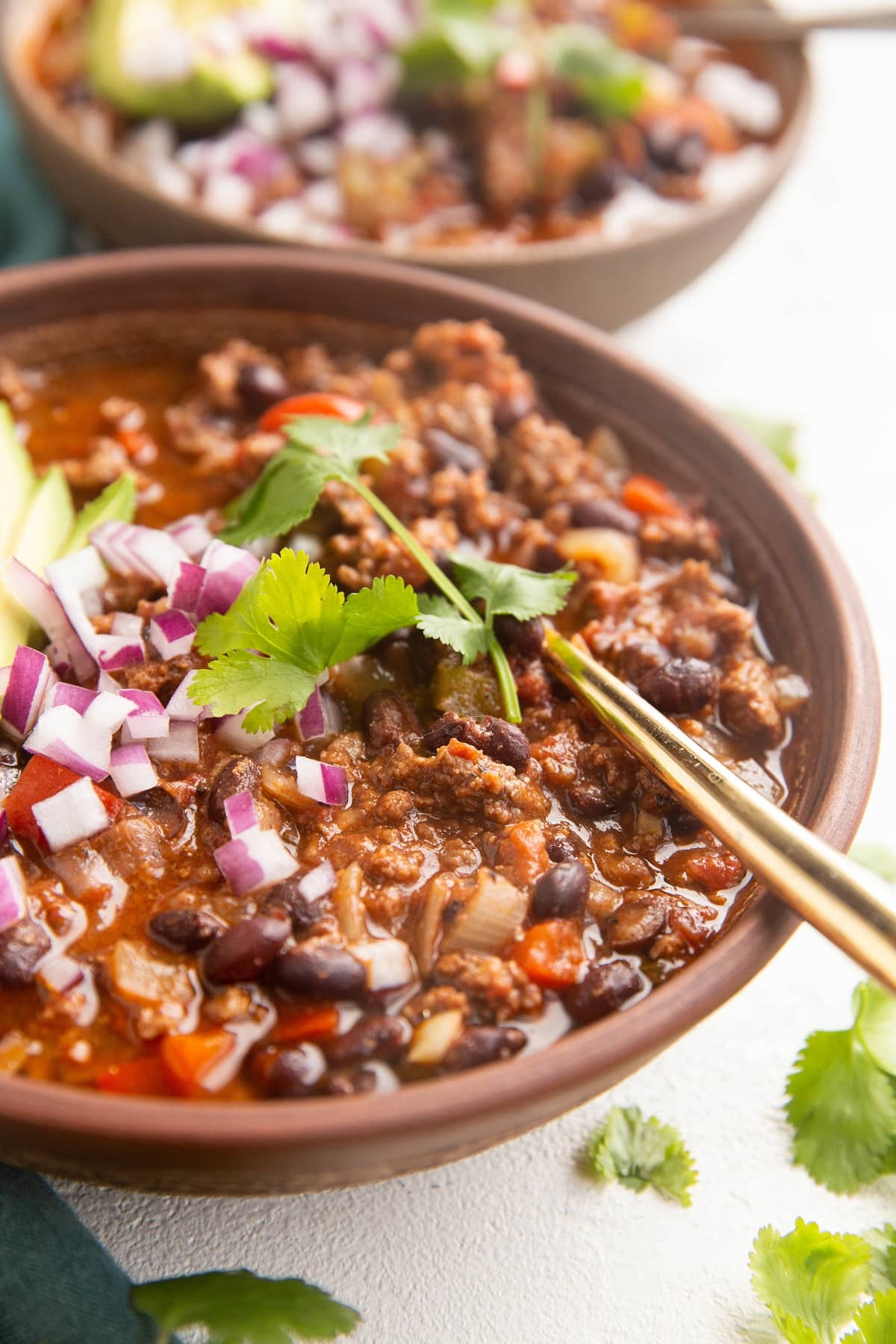 Two bowls of beef chili with a gold spoon, sprinkled with red onions and cilantro.