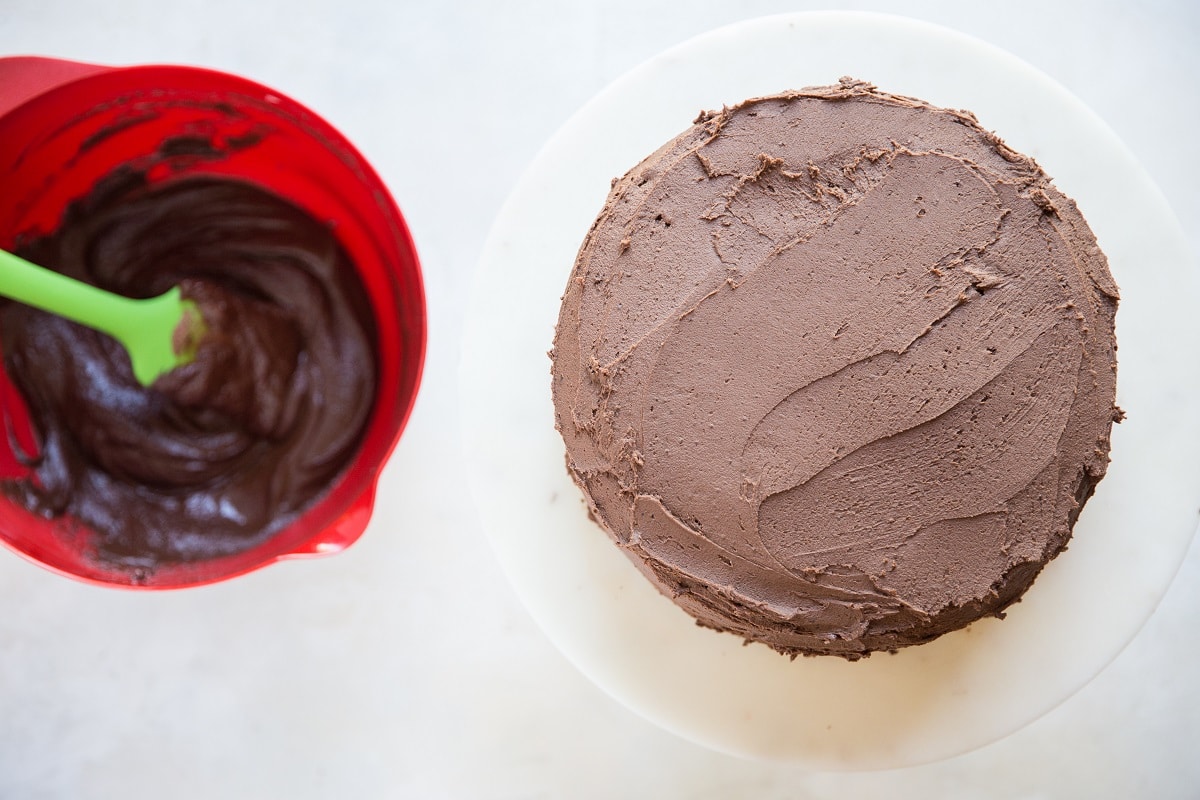 Chocolate cake on a cake stand, smothered in chocolate buttercream frosting with a bowl of ganache to the side.