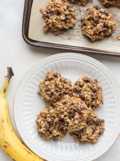 White plate stacked with healthy banana oat cookies with a ripe banana to the side and the baking sheet of the rest of the cookies to the side.