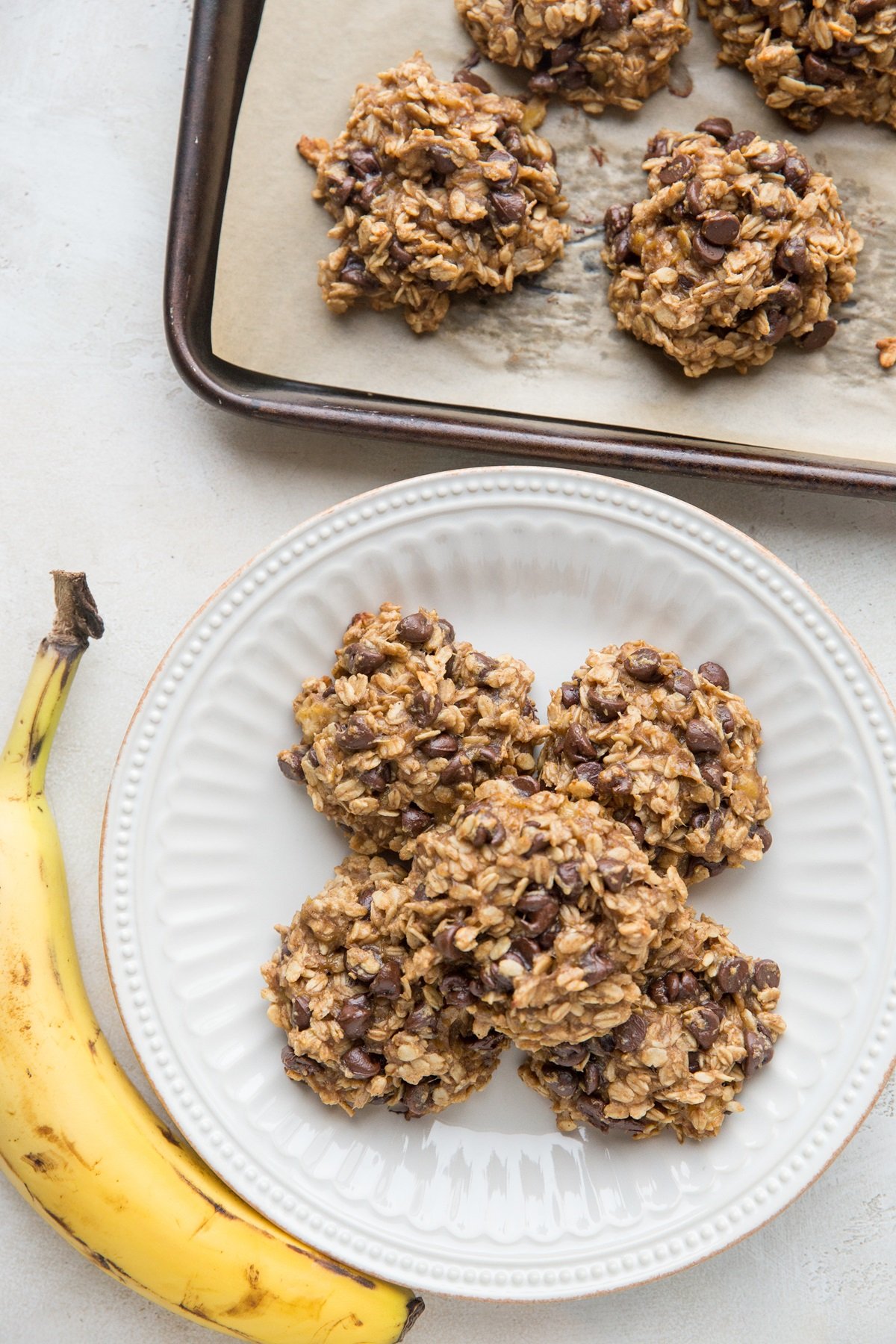 White plate stacked with healthy banana oat cookies with a ripe banana to the side and the baking sheet of the rest of the cookies to the side.