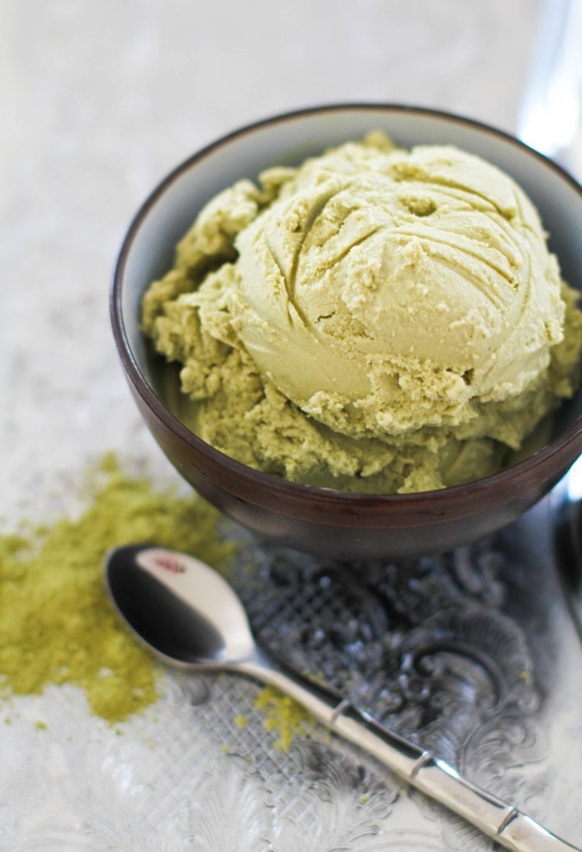 Scoop of green tea ice cream in a bowl with a spoon to the side and a sprinkle of matcha powder to the side.