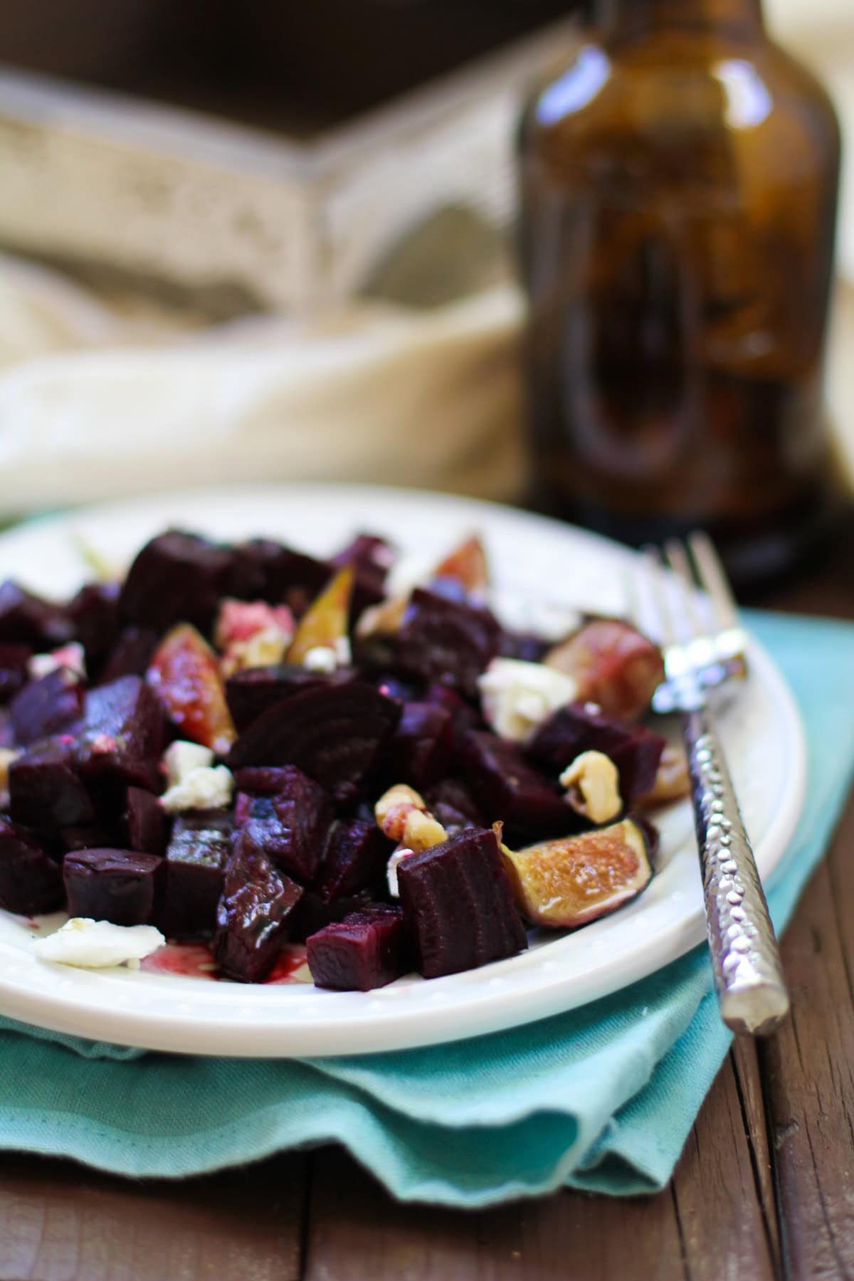 Roasted beets and figs on a white plate with walnuts and balsamic drizzle on top of a blue napkin with a fork for serving.