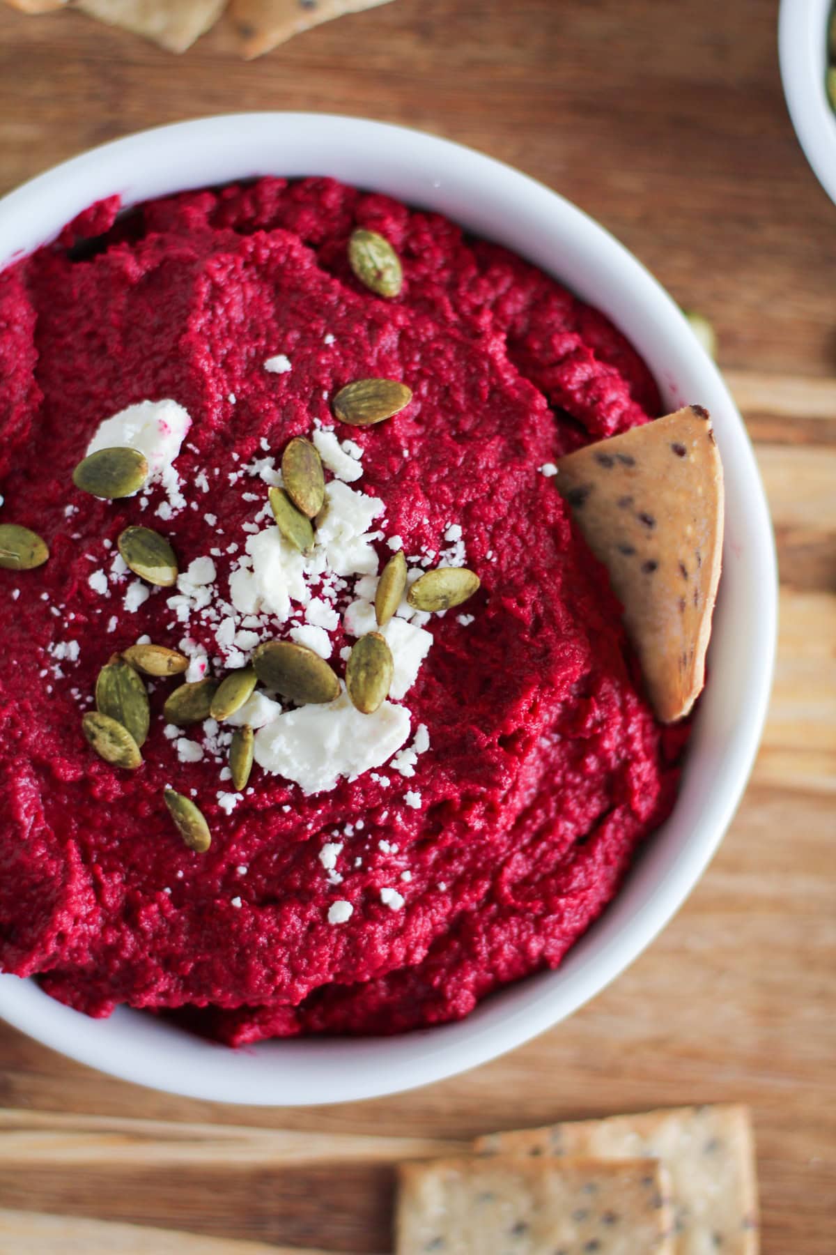 White bowl full of vibrant red beet hummus sprinkled with feta cheese and pumpkin seeds with crackers all around for serving.