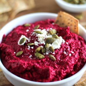 White bowl full of beet hummus with a vibrant red color. Topped with crumbled feta cheese, pumpkin seeds and crackers around for serving.