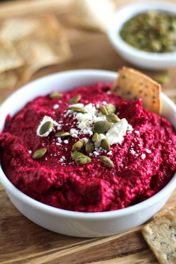 White bowl full of beet hummus with a vibrant red color. Topped with crumbled feta cheese, pumpkin seeds and crackers around for serving.