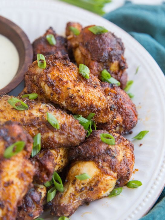 Big white plate with a pile of dry rub air fried chicken wings sprinkled with green onions with a dark blue napkin to the side.