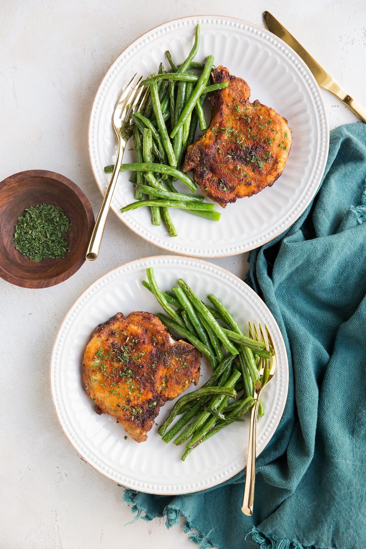 Two white plates of pork chops with air fryer green beans to the side. Blue napkin and gold forks.