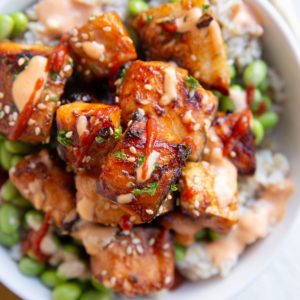 Air fryer salmon bites in a white bowl on top of steamed brown rice and edamame. A gold fork and napkin to the side.