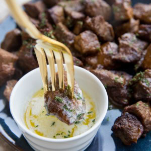 Blue plate full of air fryer steak bites with a small bowl of melted butter and a fork dipping a steak bite into the butter.