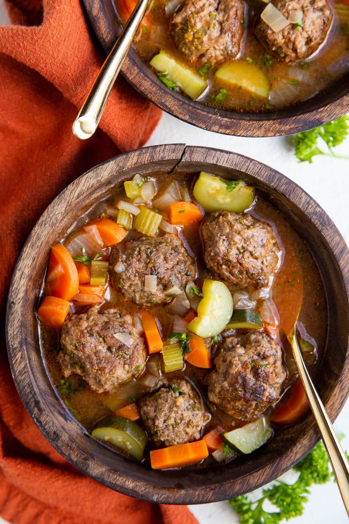 Two large wooden bowls of meatball soup with a red napkin to the side and fresh parsley.