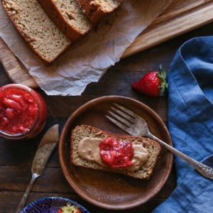 a slice of Almond Butter Banana Bread on a wooden plate with almond butter and strawberry jam spread on top with the rest of the loaf cut into slices on the side and a small bowl of fresh strawberries.