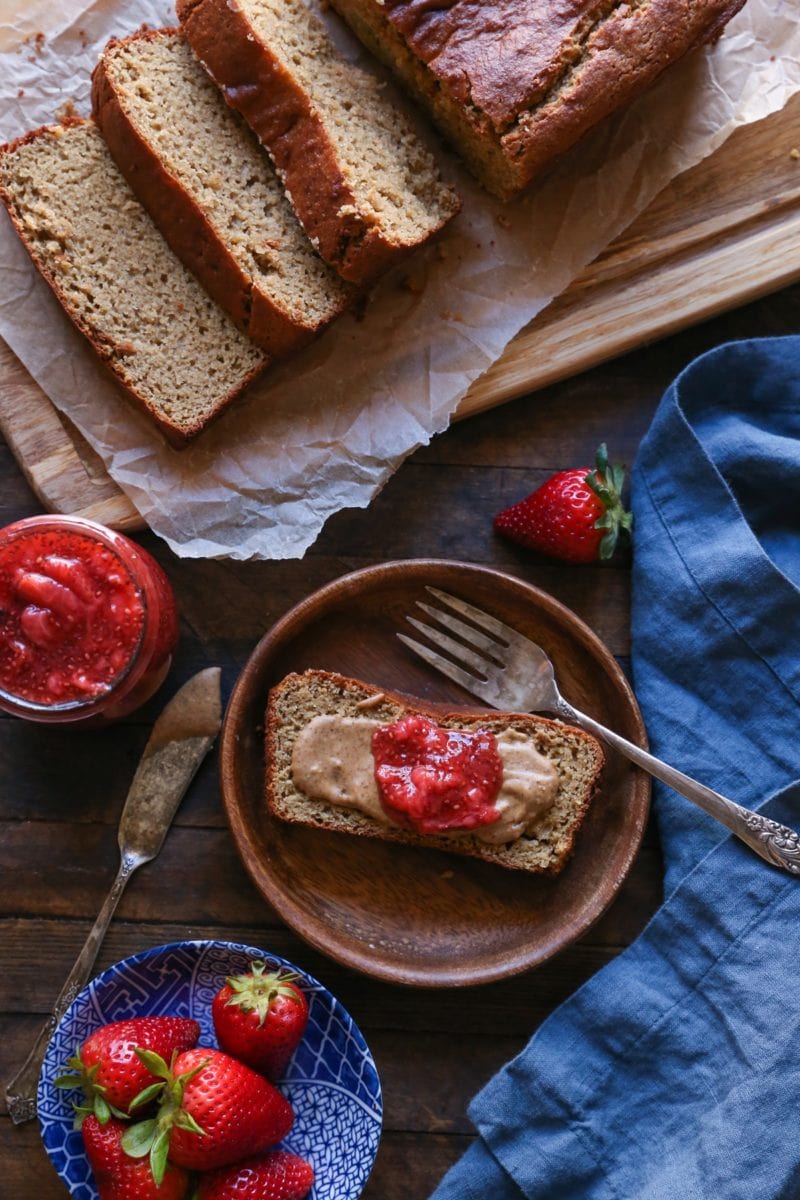 a slice of Almond Butter Banana Bread on a wooden plate with almond butter and strawberry jam spread on top with the rest of the loaf cut into slices on the side and a small bowl of fresh strawberries.