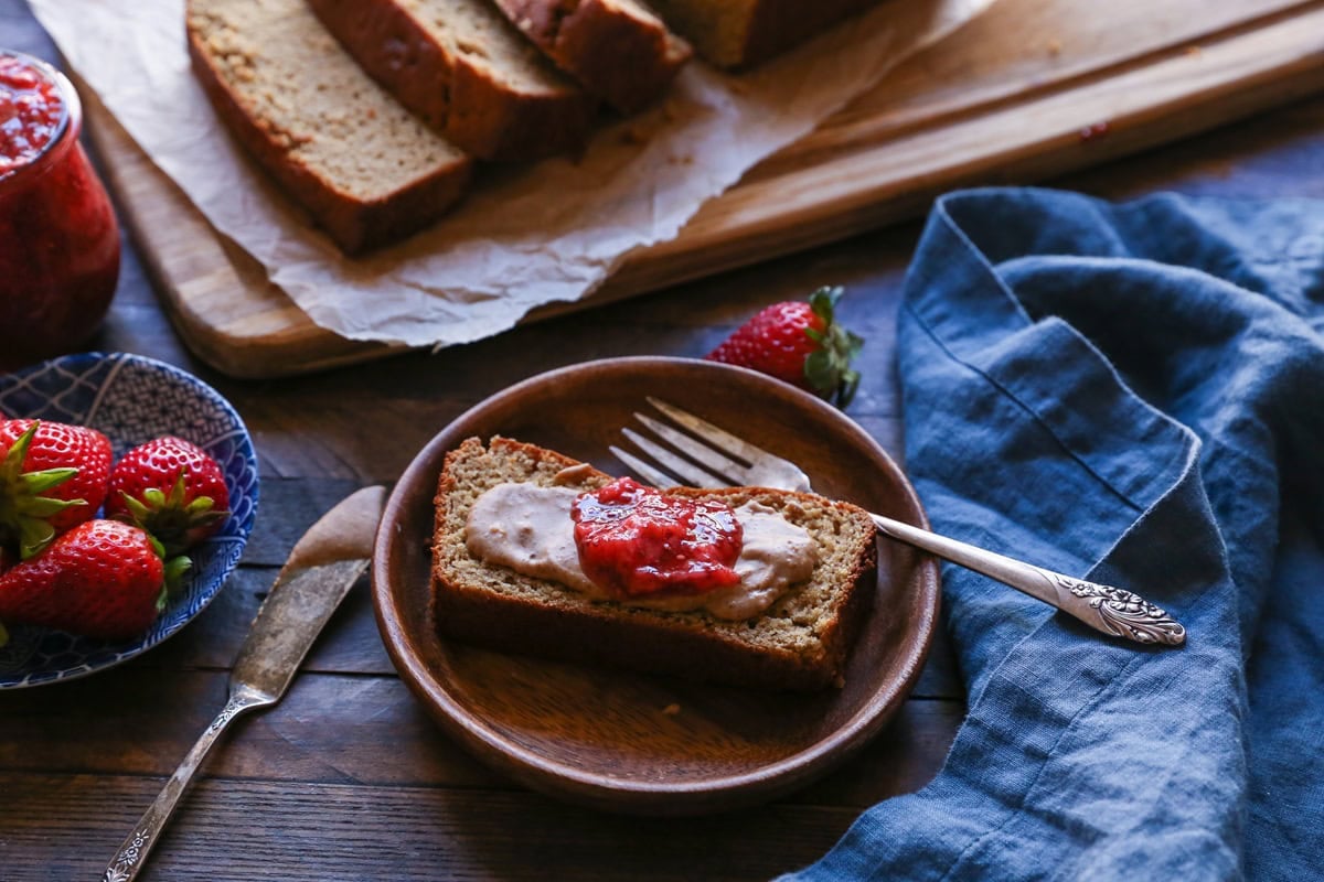 Slice of almond butter banana bread on a wooden plate with a smear of almond butter and chia seed jam on top. Fresh strawberries and a blue napkin to the side.
