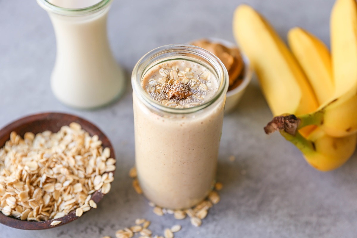 Almond Butter Protein Smoothie in a glass with a bowl of oats to the side and fresh bananas to the side.