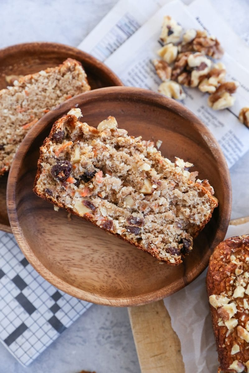 Piece of carrot cake banana bread on a wooden plate with the rest of the loaf to the side.