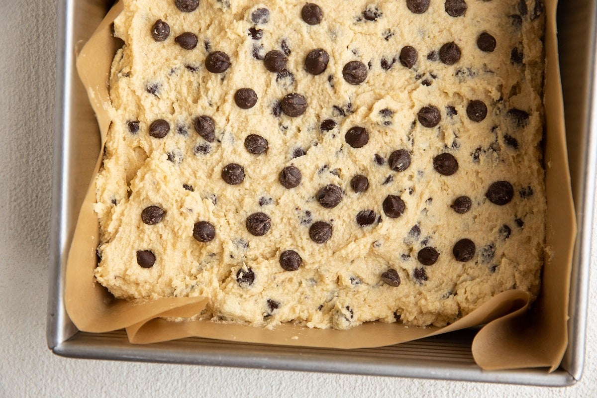 8-inch square pan lined with parchment paper with almond flour cookie dough spread into an even layer to make bars, ready to go into the oven.