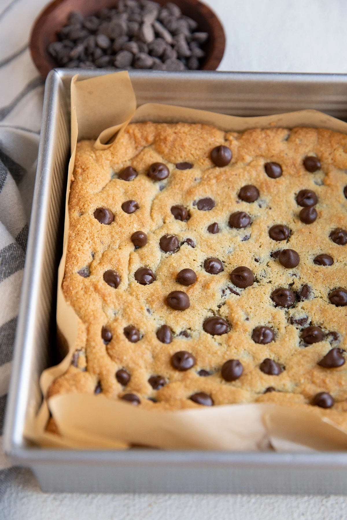 Baking pan with cookie bars inside, fresh out of the oven. A bowl of chocolate chips and a napkin to the side.