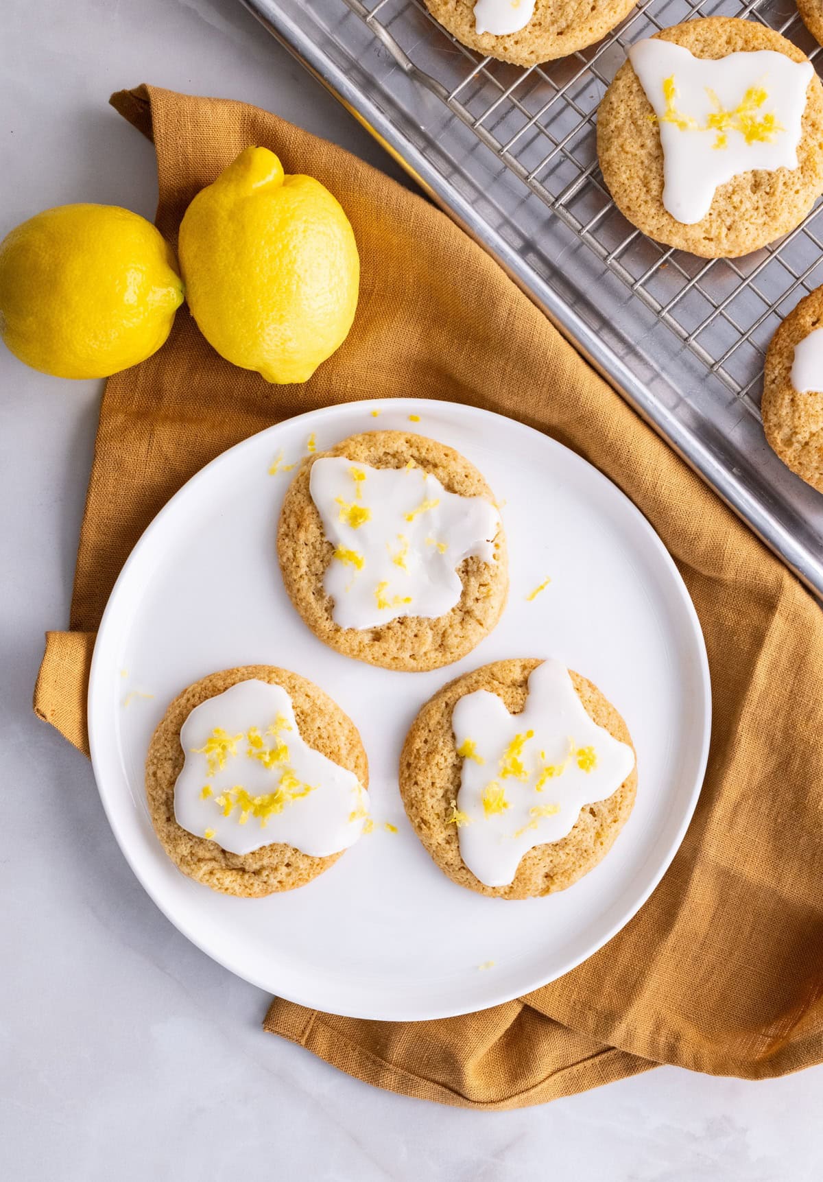 White plate of iced lemon cookies with a cookie sheet of cookies in the background.