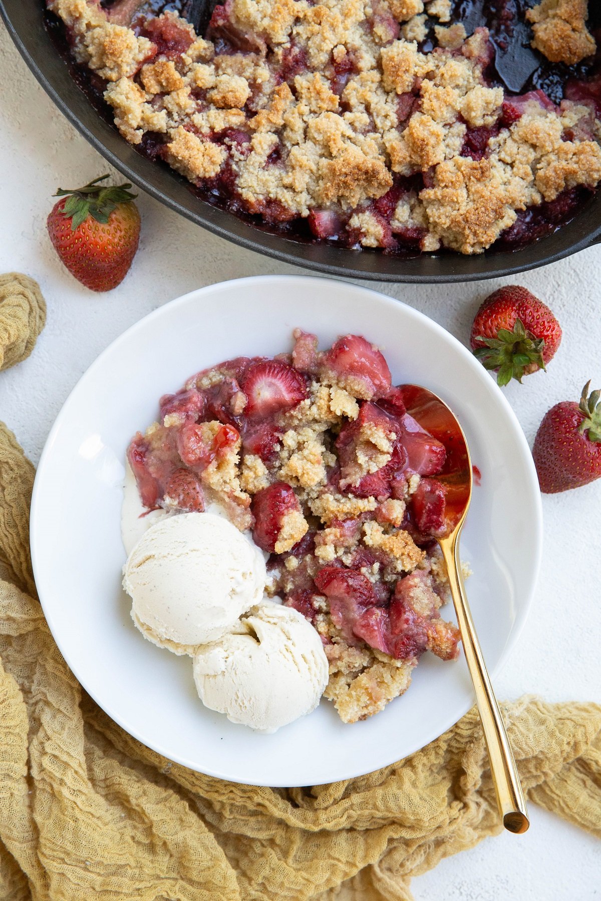 Almond flour strawberry crumble in a white bowl, ready to serve. A golden spoon and napkin to the side with fresh strawberries scattered around.
