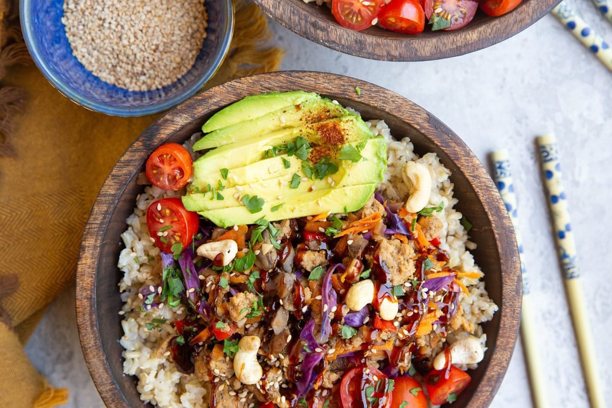 Asian ground turkey bowls in large wooden bowls with chop sticks to the side. Fresh vegetables like cabbage, carrots, and bell peppers mixed in and served with sliced avocado.