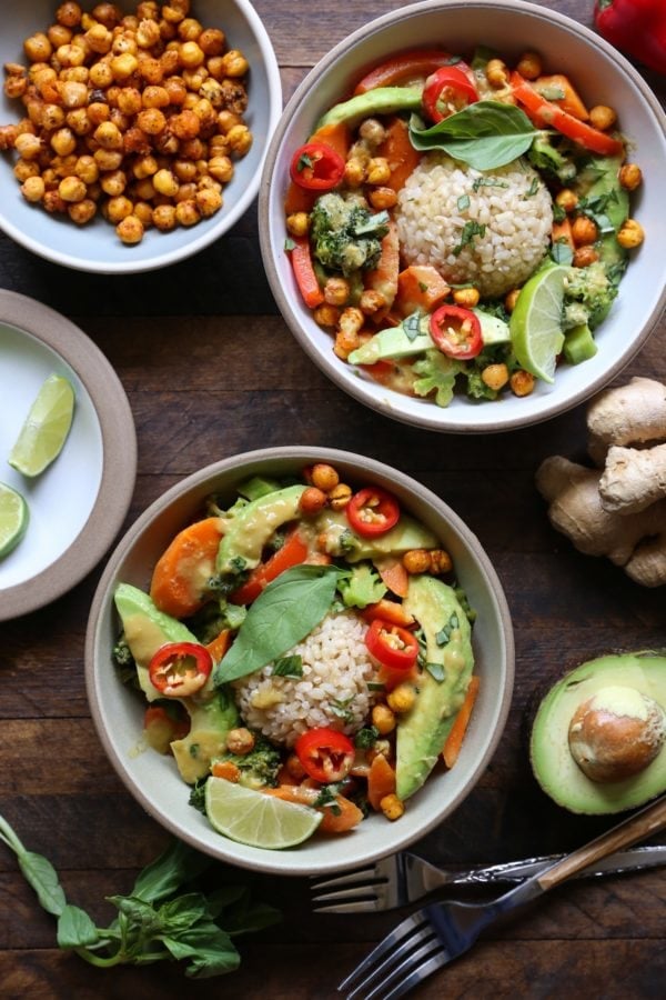 Two bowls of Thai green curry with vegetables and brown rice with forks and fresh basil to the side.