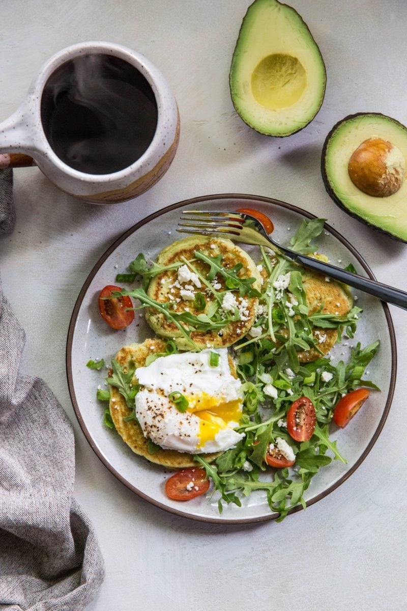 Three savory avocado pancakes on a plate with a poached egg on top and a side salad and a mug of coffee.