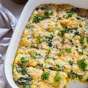 A white baking dish holds a spinach, cheese, and bacon and egg casserole, cut into square pieces and garnished with fresh parsley. Plates, forks, and a striped towel are nearby on a light-colored surface.