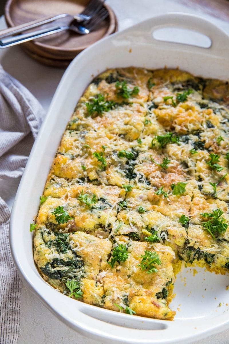 A white baking dish holds a spinach, cheese, and bacon and egg casserole, cut into square pieces and garnished with fresh parsley. Plates, forks, and a striped towel are nearby on a light-colored surface.