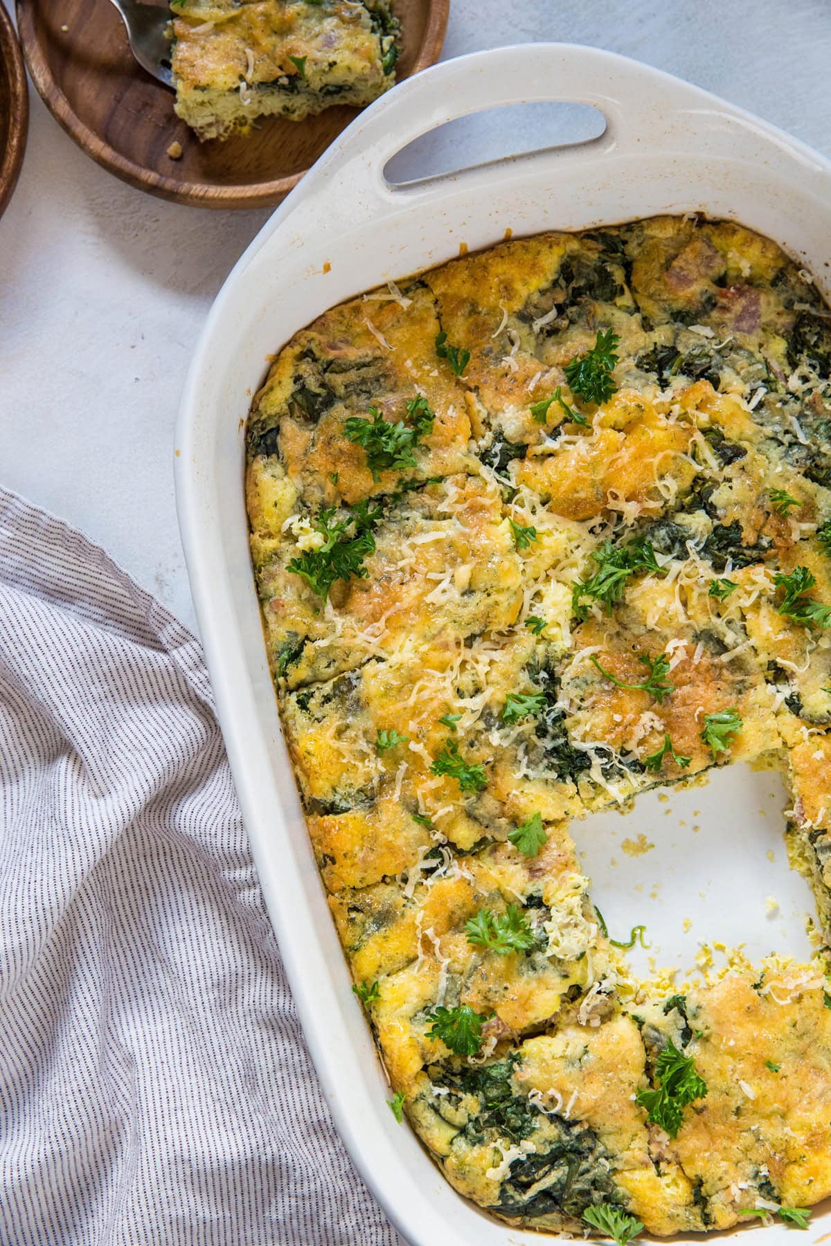 A bacon and egg casserole with greens and cheese, topped with fresh parsley, sits in a white baking dish. A square piece has been removed, revealing the fluffy, golden interior. A linen napkin and a plated serving are nearby.