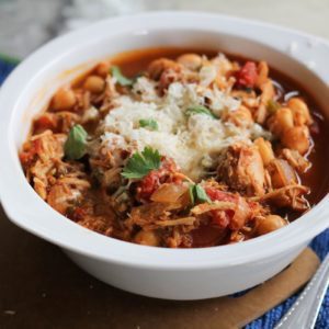 Barbecue Pulled Chicken Chili in a white bowl with cilantro and cheese on top. A spoon and a blue napkin to the side.