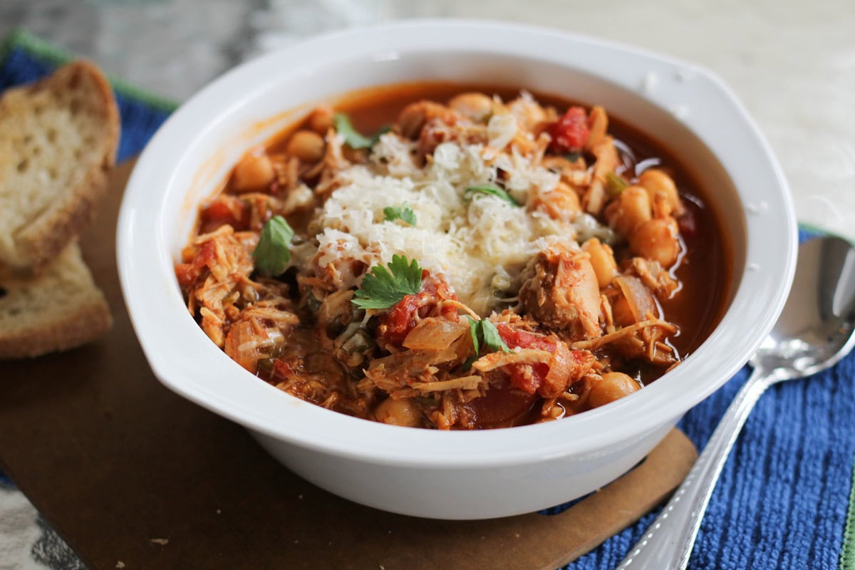 Barbecue Pulled Chicken Chili in a white bowl with cilantro and cheese on top. A spoon and a blue napkin to the side.