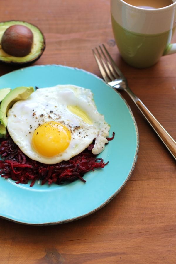Beet Hash Browns on a blue plate with an egg and avocado on top. The plate sits on a wooden table with a cup of coffee and a fork to the side.