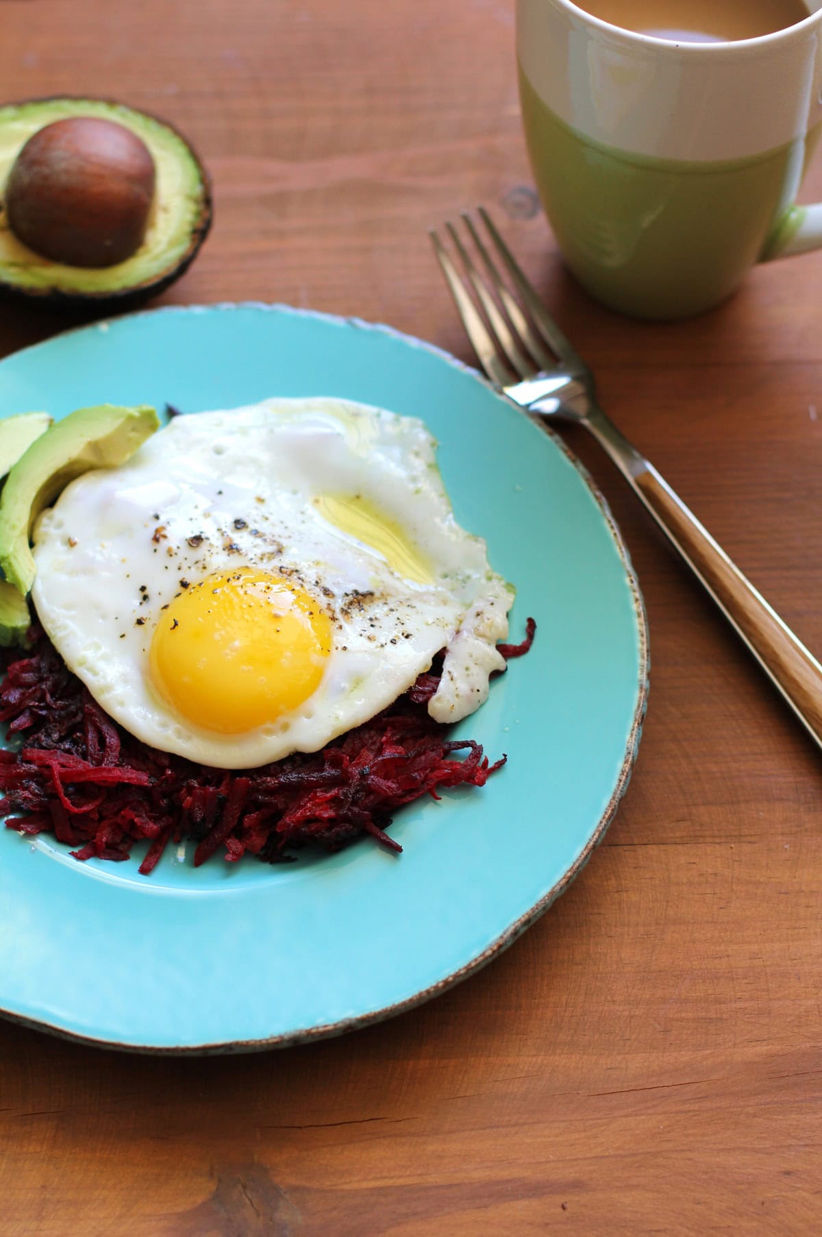Beet Hash Browns on a blue plate with an egg and avocado on top. The plate sits on a wooden table with a cup of coffee and a fork to the side.