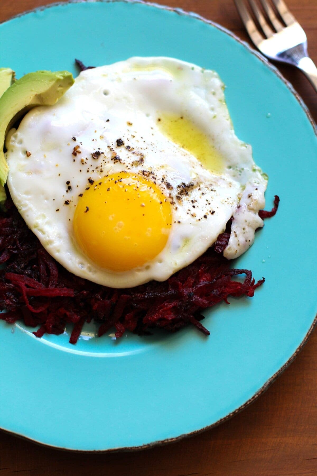 Beet Hash Browns on a blue plate with a sunny side up egg and avocado on top. Ready to eat.