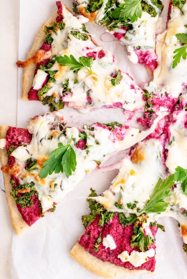 Pizza cut into slices on a sheet of parchment paper with beet pesto sauce, kale, and goat cheese.