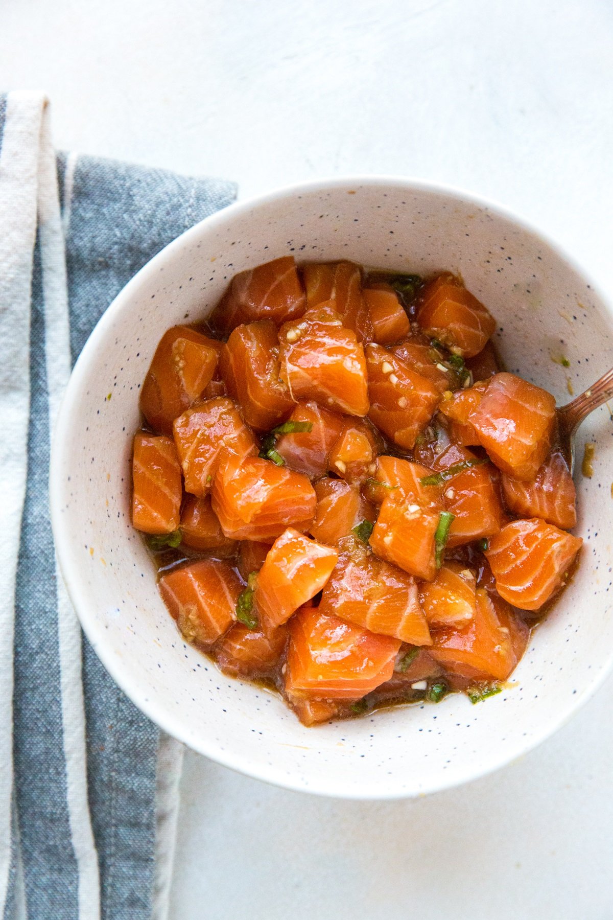 Bowl of salmon poke with spoon and blue napkin on white surface
