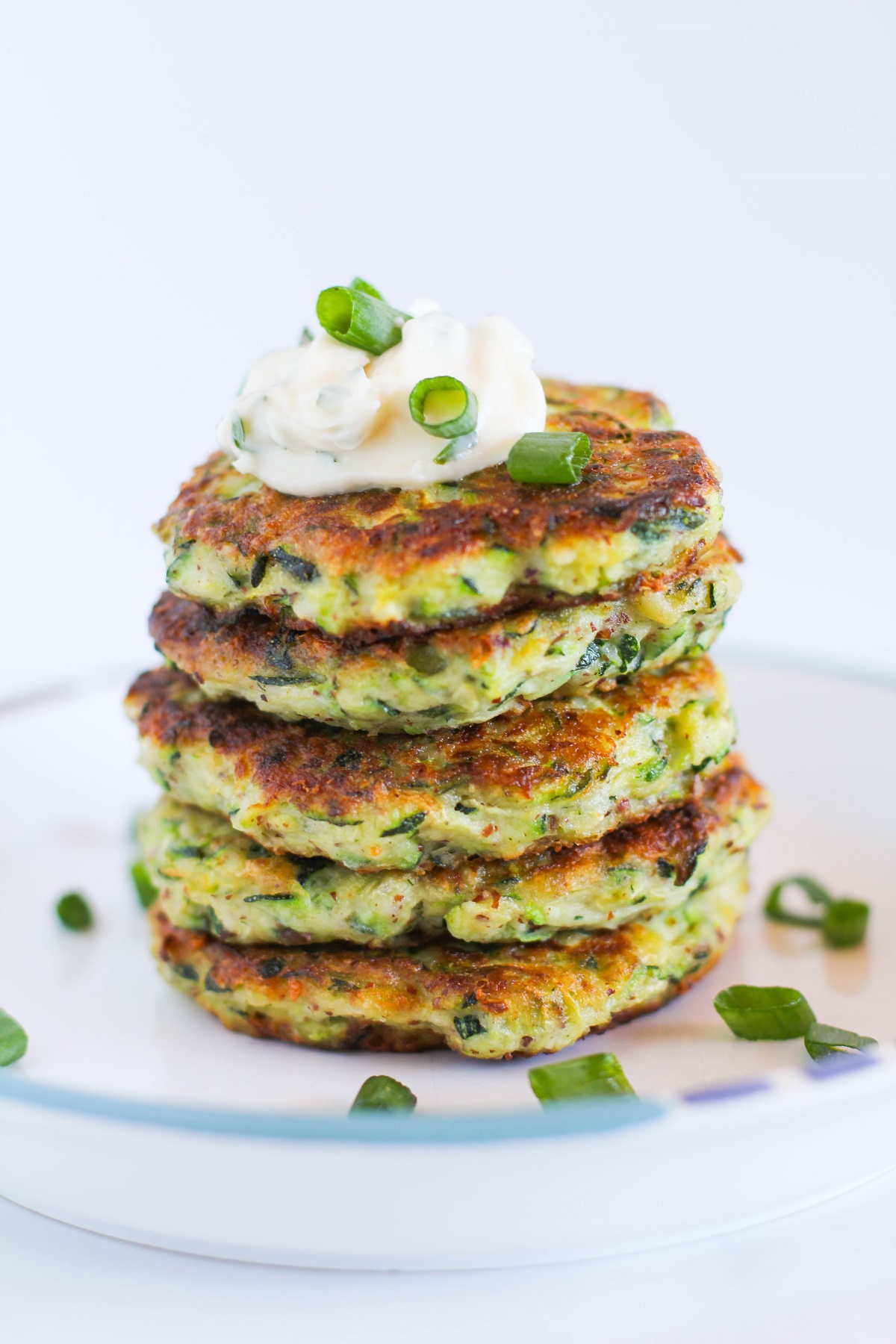 Stack of zucchini fritters on a plate with a dollop of aioli on top and chopped green onions, ready to serve.