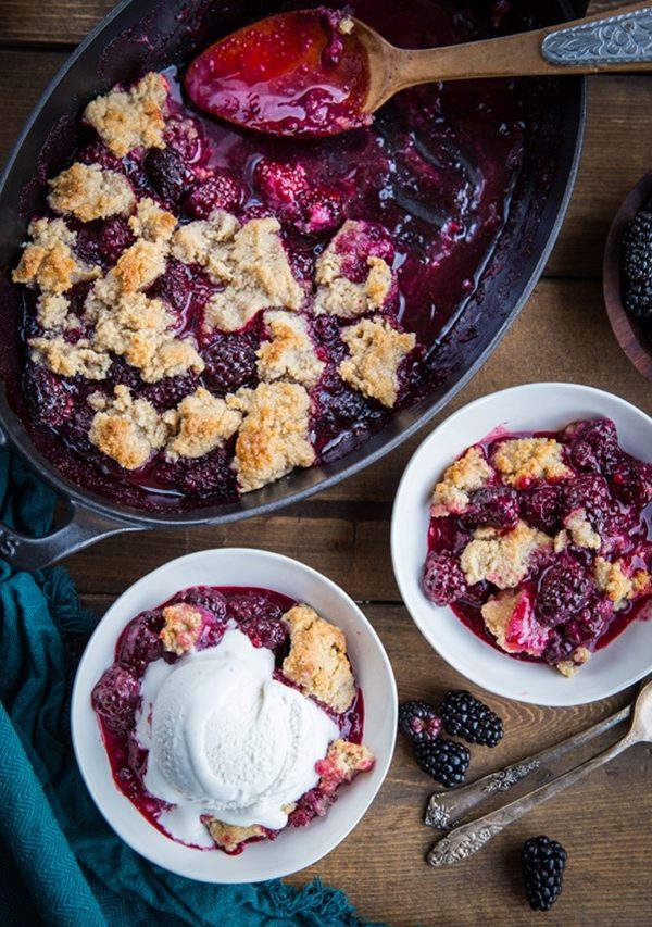 Paleo Blackberry Cobbler in two bowls, one with a scoop of ice cream on top, and the rest of the cobbler in a cast iron baking dish with fresh blackberries to the side, ready to serve.