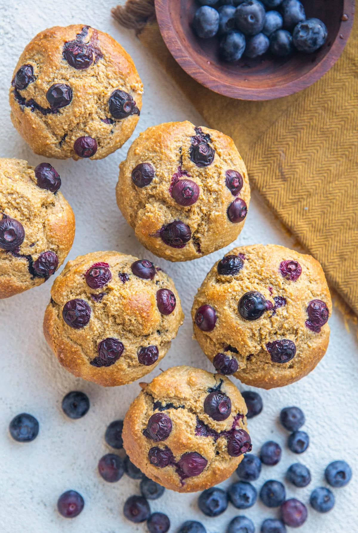 Six blueberry banana muffins on a white background with a golden napkin and fresh blueberries scattered about.
