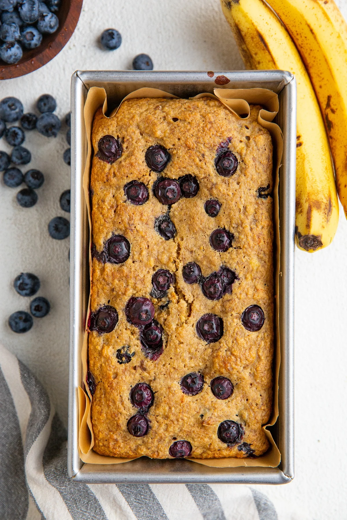 Loaf of blueberry oatmeal bread in a bread loaf pan with ripe bananas and blueberries to the side.