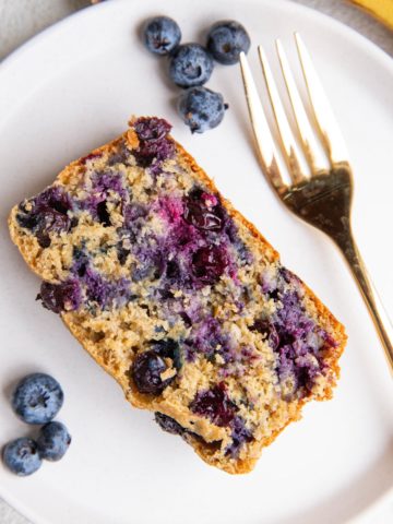 Slice of blueberry banana oatmeal bread on a white plate with fresh blueberries and banana to the side and a gold fork.