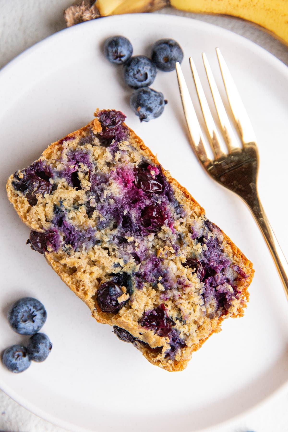 Slice of blueberry banana oatmeal bread on a white plate with fresh blueberries and banana to the side and a gold fork.