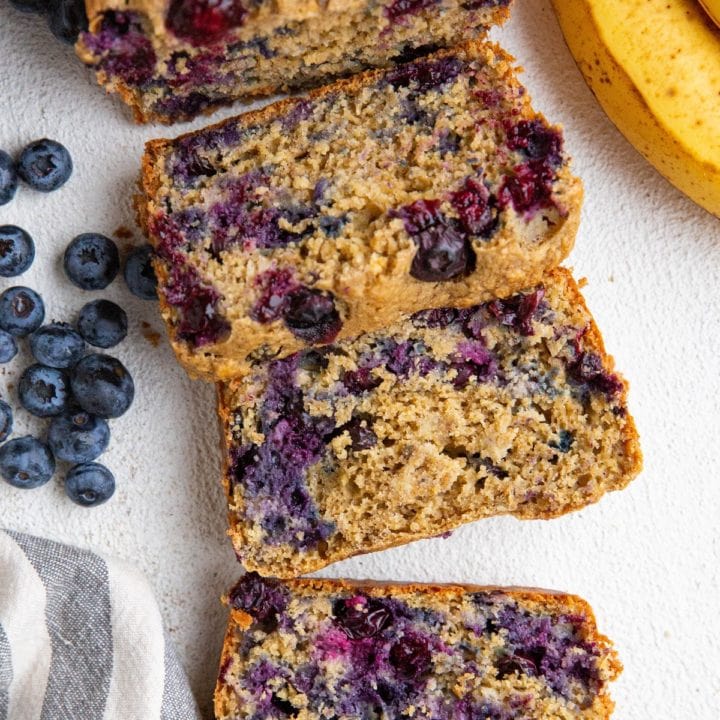 Loaf of banana oatmeal bread cut into slices with ripe bananas and blueberries to the side.