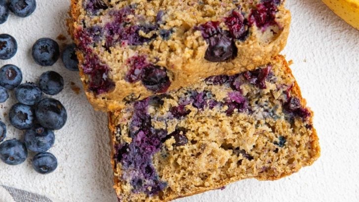 Loaf of banana oatmeal bread cut into slices with ripe bananas and blueberries to the side.