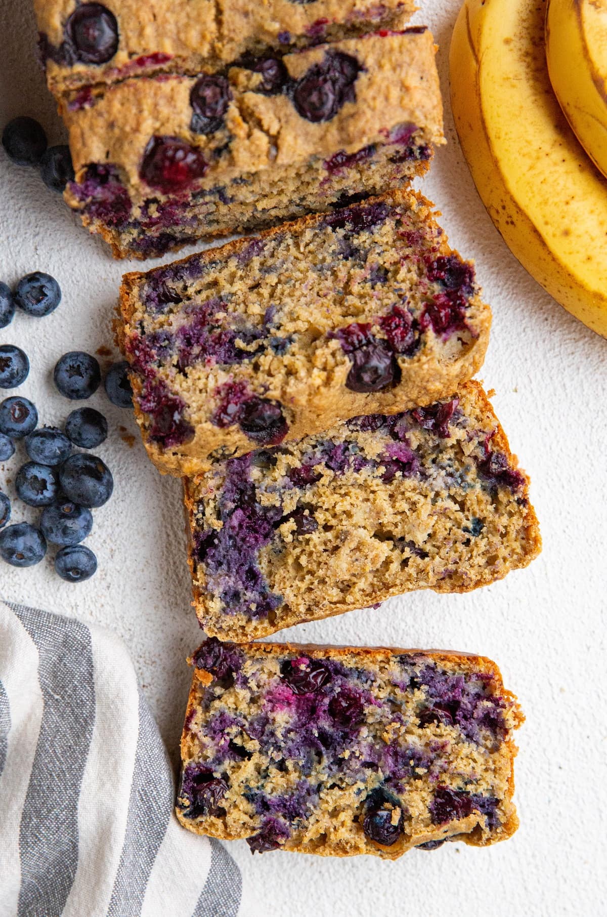 Loaf of banana oatmeal bread cut into slices with ripe bananas and blueberries to the side.