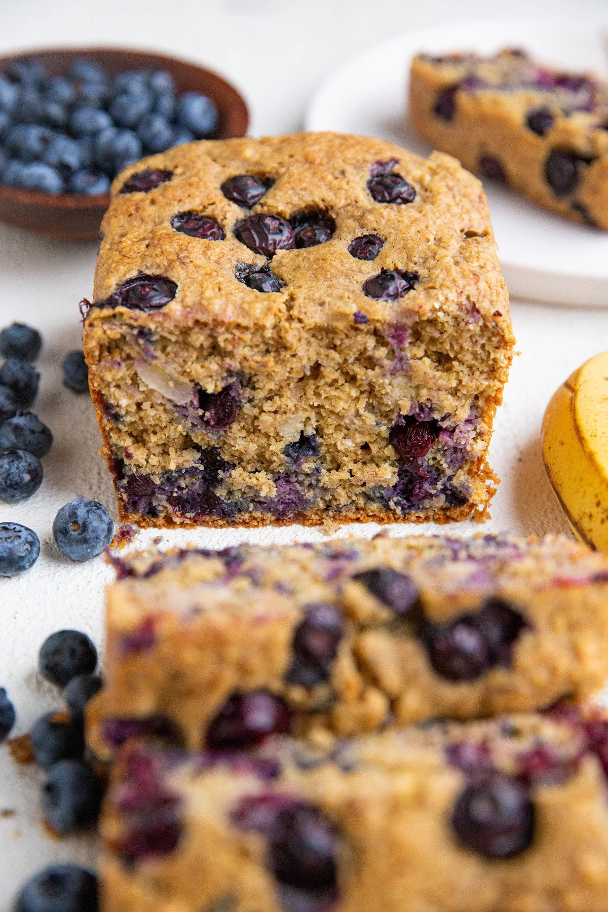 Loaf of blueberry banana bread cut into slices with fresh berries and bananas to the side.