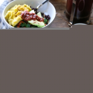 Two breakfast bowls with sweet potatoes, greens, eggs, avocado, salsa, and cheddar cheese on a wooden table with a French press full of coffee.