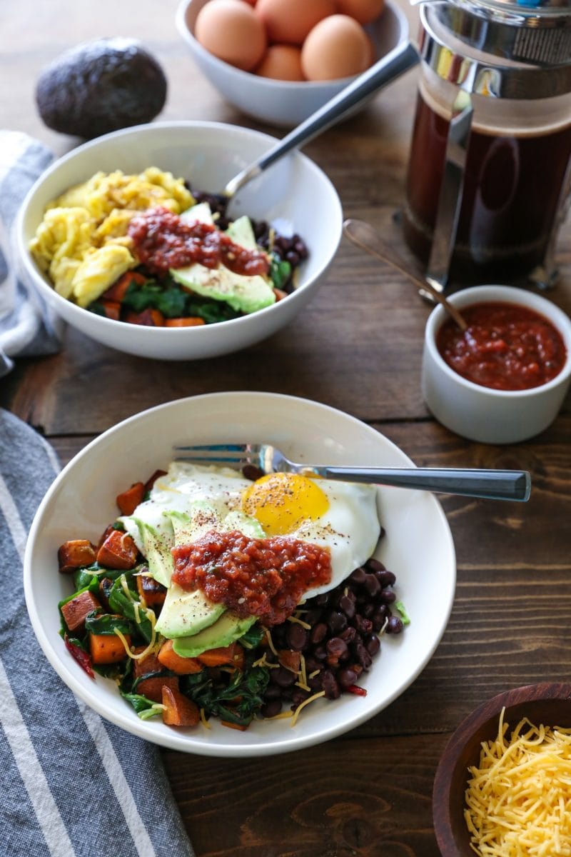 Two breakfast bowls with sweet potatoes, greens, eggs, avocado, salsa, and cheddar cheese on a wooden table with a French press full of coffee.
